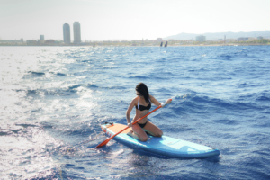 A woman kneeling on a blue paddleboard in the shimmering Mediterranean Sea off the coast of Barcelona. In the background, the iconic Olympic towers (Hotel Arts and Torre Mapfre) are visible against a bright, sun-drenched skyline. This represents the active leisure portion of a luxury sailing and water sports tour.