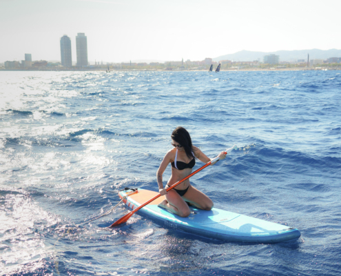 A woman kneeling on a blue paddleboard in the shimmering Mediterranean Sea off the coast of Barcelona. In the background, the iconic Olympic towers (Hotel Arts and Torre Mapfre) are visible against a bright, sun-drenched skyline. This represents the active leisure portion of a luxury sailing and water sports tour.
